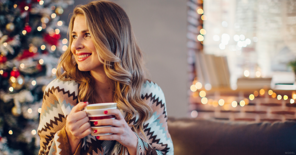 With a Christmas tree and lights in the background, a smiling woman has a blanket wrapped around her and holds a mug.