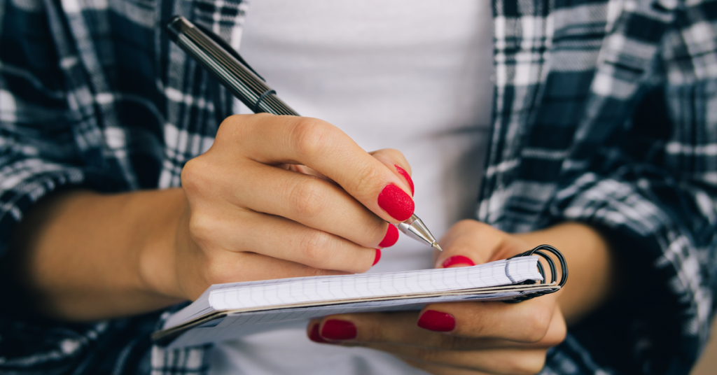 Close-up of hands with red fingernails using a pen to write on a spiral-bound notebook.