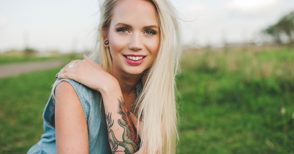 Blonde woman kneels in a grassy field with her hand on her shoulder, showing the colorful tattoo on her forearm.