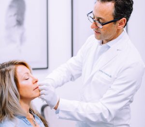 Standing and wearing a white lab coat, Dr. Rockmore examines patient\'s face.