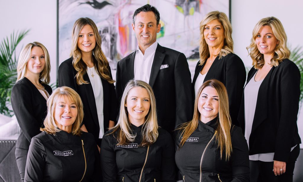 The Rockmore Plastic Surgery team, wearing black suits and white shirts or black uniforms, stands in the office lobby.