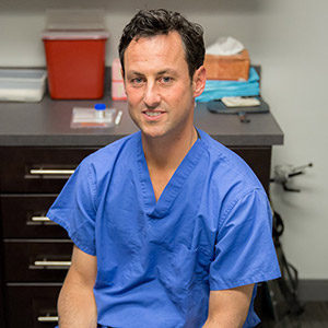 In a blue scrub top, Dr. Rockmore sits in front of a cabinet with medical supplies, looking directly at the camera.