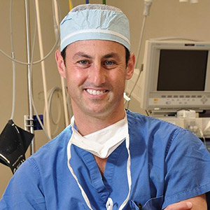 Dr. Rockmore smiles at the camera, wearing a surgical cap, a partially tied mask, and blue scrubs.