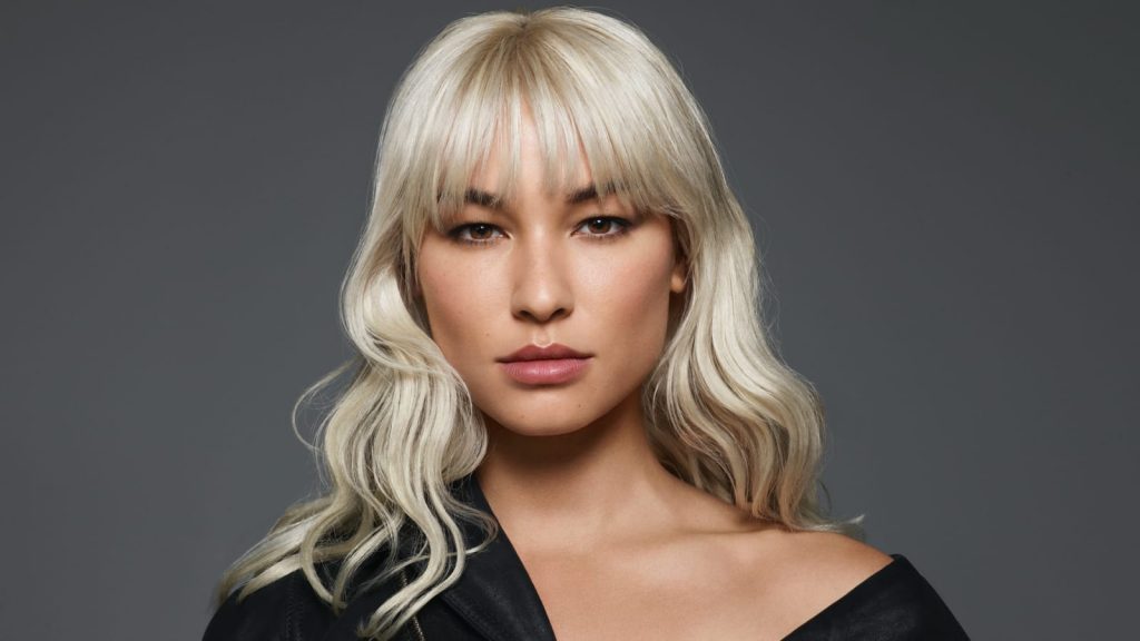 Close-up of a striking woman with long platinum blond hair and brown eyes gazes directly at the camera.