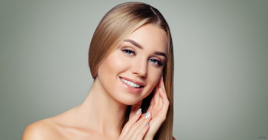A blonde woman smiles softly while gently touching her face. (MODEL)